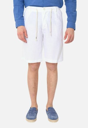 Uomo che indossa pantaloncini bianchi al ginocchio con coulisse, una camicia blu a maniche lunghe e scarpe slip-on blu, in piedi.