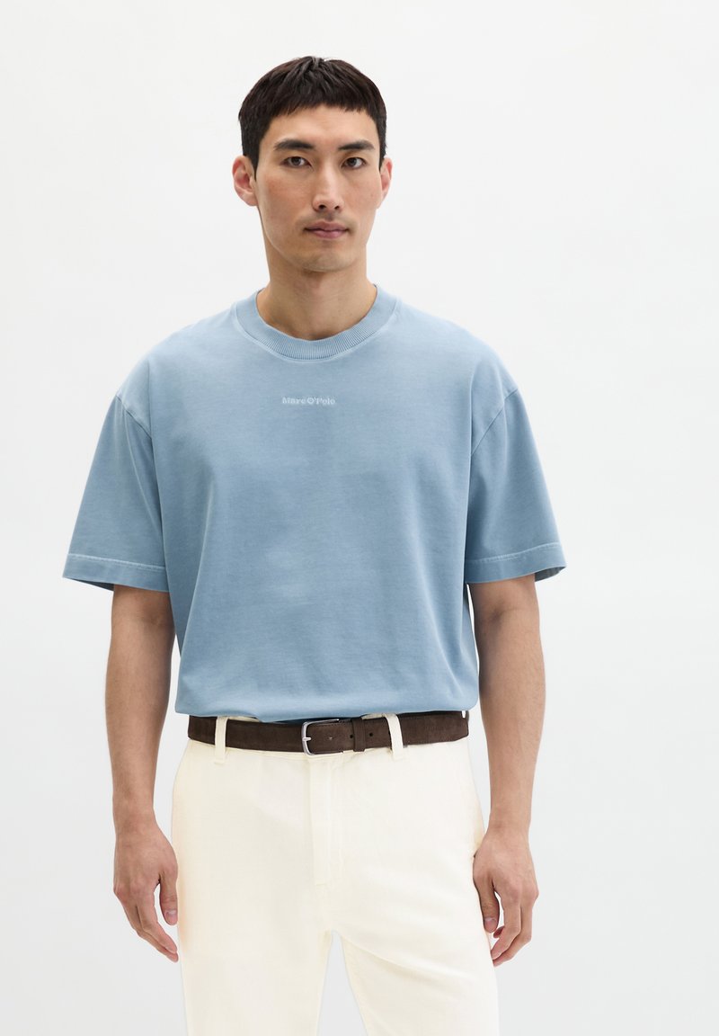 Homme portant une chemise à manches courtes bleu clair rentrée dans un pantalon crème avec une ceinture marron foncé, debout devant un fond blanc uni.