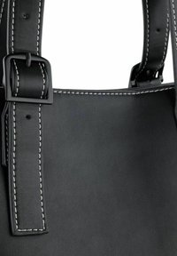 Detail van een zwarte leren tas met een gestructureerde oppervlakte, contrasterende witte stiksels en een zwarte metalen gesp op een verstelbare schouderband.