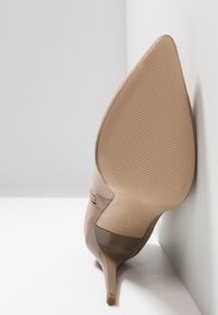 Chaussure à talon haut beige à bout pointu avec semelle striée et talon aigu fin reposant contre un mur blanc et un sol blanc.