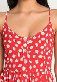 Vestido rojo con estampado de margaritas blancas, tejido ligero, tirantes ajustables y detalles de botones en la parte delantera. Sección de falda acampanada.