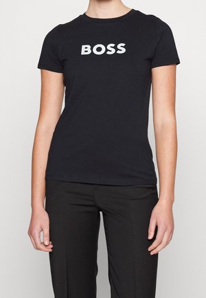 Persoon draagt een nauwsluitend zwart T-shirt met "BOSS" in witte letters centraal op de borst, gecombineerd met een zwarte broek.