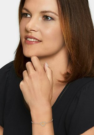 Silberarmband mit einem herzförmigen Anhänger, verziert mit goldenen Akzenten und eingelassenen Steinen. Die Trägerin hat eine nachdenkliche Pose und schulterlanges Haar.
