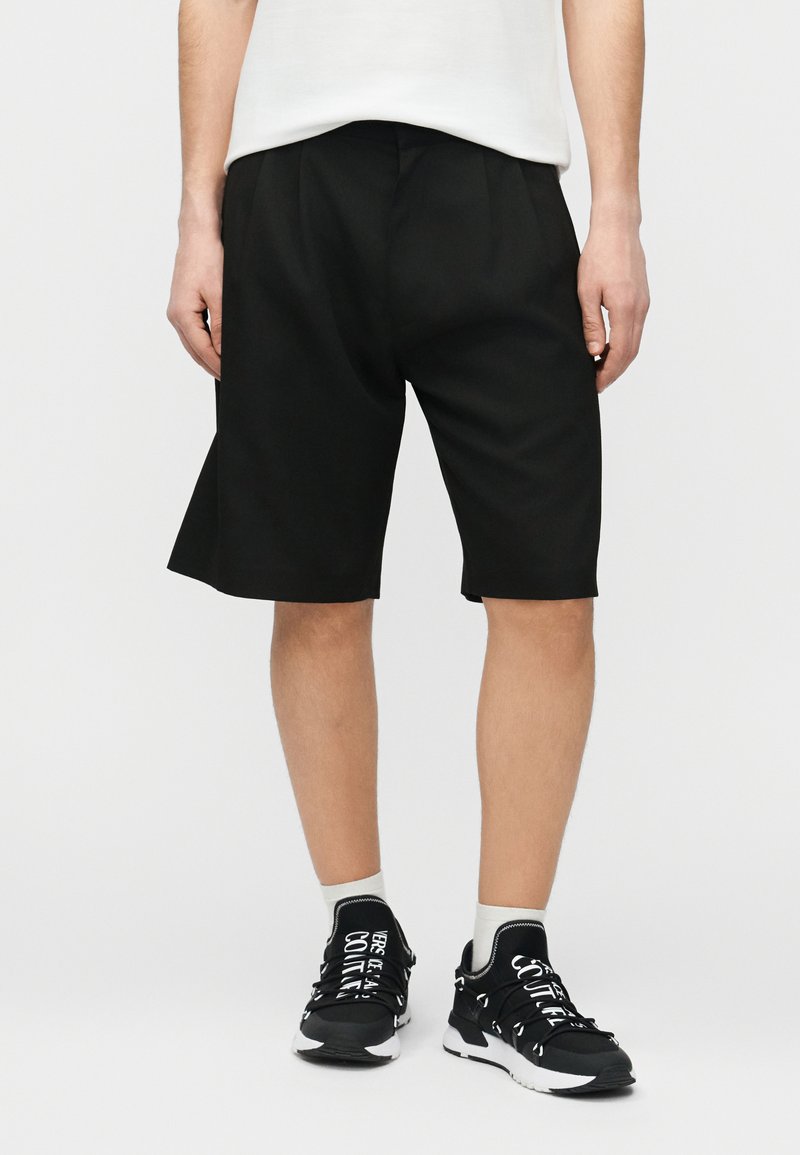 Άνδρας που φοράει μαύρα σορτς μέχρι το γόνατο, λευκές κάλτσες και μαύρα αθλητικά παπούτσια με λευκό κείμενο, στέκεται μπροστά σε απλό φωτεινό φόντο.