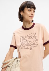 Κοντομάνικο, ανοιχτό ροζ βαμβακερό μπλουζάκι με σκουροκόκκινη ριγωτή γιακά. Διαθέτει κεντητό γραφικό με σχέδια φυτών και ζώων.