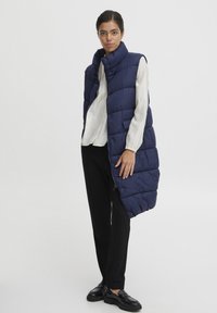 Vest lung din material căptușit, de culoare bleumarin, cu guler înalt, care prezintă închidere cu nasturi și fermoar. Este purtat peste un top de culoare deschisă și pantaloni negri.