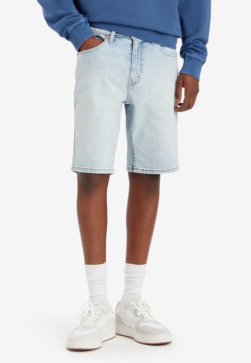 Shorts en denim bleu clair avec une coupe droite, cinq poches et passants pour ceinture, associés à des baskets blanches et des chaussettes. Design décontracté.