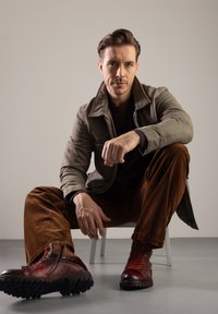 Hnědé manšestrové kalhoty, šedá bunda, tmavá košile a červené poškozené boty s texturovanými podrážkami. Subjekt sedící na židli s neutrálním pozadím.