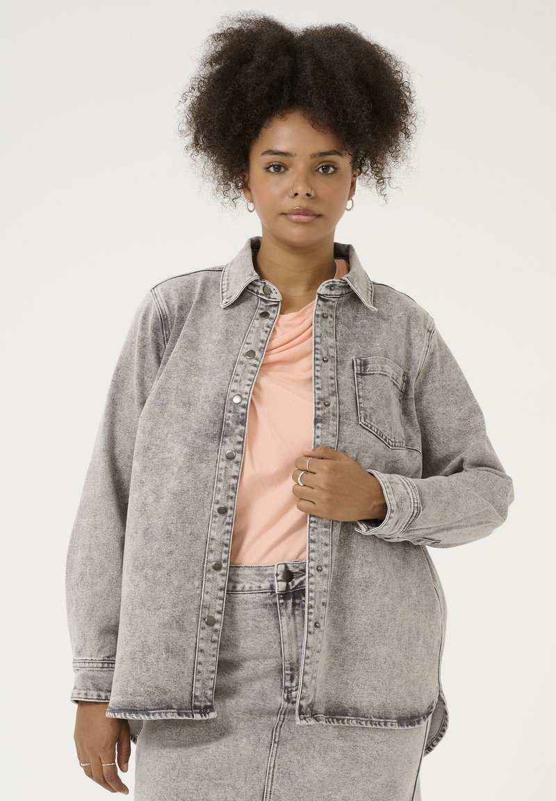 Grå denimskjorte med knaplukning foran, krave og brystlomme. Båret over en lys pink top, suppleret af en matchende pink nederdel.