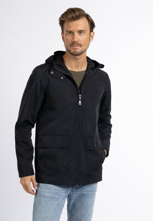 Mann trägt eine schwarze Kapuzenjacke mit Reißverschluss und Fronttaschen, kombiniert mit hellblauen Jeans, stehend vor einem schlichten weißen Hintergrund.