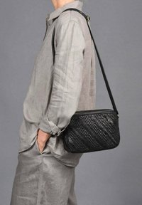 Bolso cruzado de cuero trenzado negro con cierre de cremallera y correa ajustable, combinado con un atuendo de lino gris claro. Textura suave.