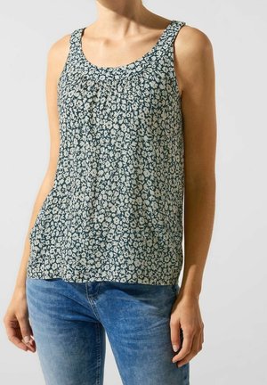 Kvinde iført en ærmeløs, mørkegrøn blomstret bluse parret med blå jeans, stående mod en ensfarvet lys baggrund.