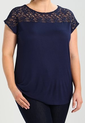 Woman wearing a navy blue short-sleeve top with floral lace detail across the yoke and shoulders, paired with dark pants.