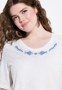 Bílé tričko s krátkým rukávem a modrým vyšíváním ryb a vinic na výstřihu. Měkká látka a volný střih. Hnědé dřevěné kroužkové náušnice.