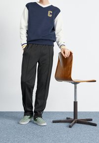 Modrá a bílá mikina s výstřihem do V a zlatým logem "C", doplněná černými kalhotami a zelenými kostkovanými nazouvacími botami, vedle dřevěné židle.