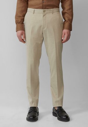 Pantalons beiges avec une texture à motif subtil, coupe droite et plis devant, associés à des chaussures noires à enfiler.