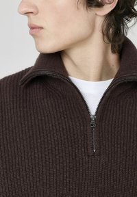 Pull en ribbed marron avec un col zippé à moitié. Présente un tissu texturé et une fermeture éclair contrastante. Porté par-dessus une chemise blanche.