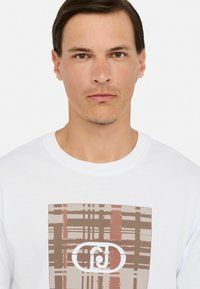 Balta T-kreka ar centrālu grafiku, kurā ir brūni un sarkani svītrainie raksti, izceltu ar apļa logotipu, īsām piedurknēm un apaļu kakla izgriezumu.