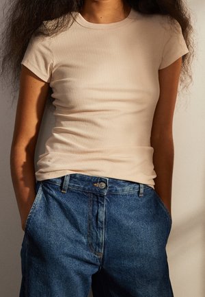 Persona con una camiseta de manga corta beige acanalada y vaqueros azules de cintura alta con las manos en los bolsillos, de pie contra una pared clara.