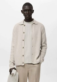 Beżowy, prążkowany kardigan zapinany na guziki, noszony na jasnobeżowej koszuli, zestawiony z beżowymi spodniami. Tkanina jest miękka z delikatnymi detalami tekstury.