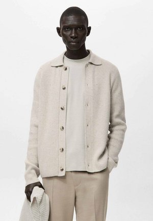 Cardigan beige côtelé avec boutons, porté sur une chemise beige clair, associé à un pantalon beige. Le tissu est doux avec des détails subtils de texture.