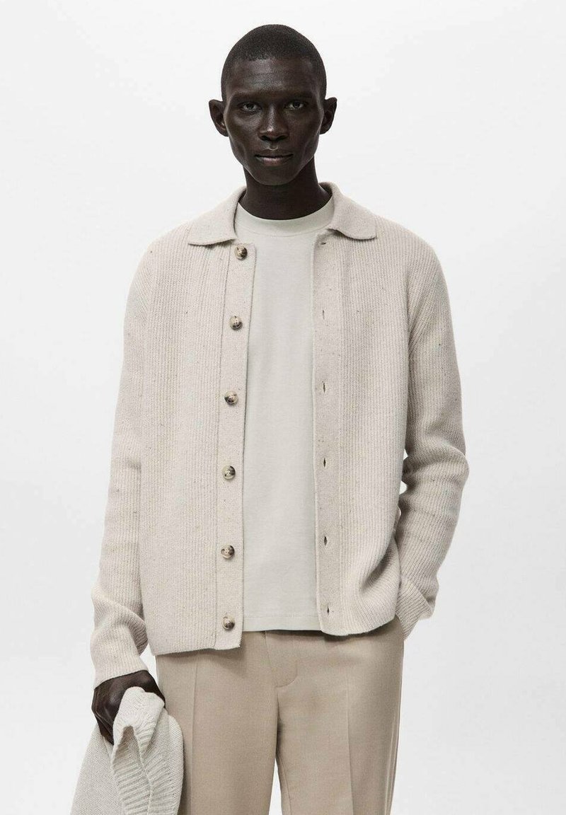 Beżowy, prążkowany kardigan zapinany na guziki, noszony na jasnobeżowej koszuli, zestawiony z beżowymi spodniami. Tkanina jest miękka z delikatnymi detalami tekstury.