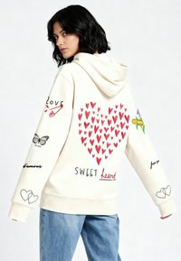 Kremasti hoodie s velikim crvenim grafičkim srcem i raznim printanim motivima, uključujući leptire i tekst, na stražnjoj strani i rukavima.