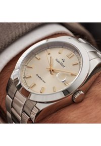 Silberne Edelstahl-Uhr mit einem cremfarbenen Zifferblatt, goldenen Stundenmarkierungen und einem Datumsfenster. Sie verfügt über eine polierte Lünette und ein strukturiertes Armband.