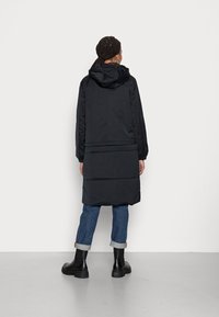 Długa czarna puchowa kurtka z kapturem, z pikowanym wzorem, elastycznymi mankietami i prostym dołem, noszona na niebieskich podwiniętych dżinsach oraz czarnych butach.