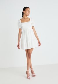 Λευκό φόρεμα με πούφ μανίκια, τετράγωνη λαιμόκοψη και στρωματοποιητή φούστα. Η λείο, υφασμάτινο υλικό αντίκειται με τα ροζ σανδάλια με λουριά.