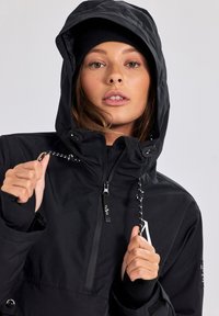 Giacca impermeabile nera con cappuccio, zip frontale e polsini elasticizzati. Presenta cordini regolabili e toppe con logo.