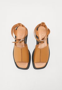 Bruna, öppna sandaler med en slät läderöverdel, i fyrkantig form och en svart plattformssula. Har ankelsnoddar och runda metalldetaljer.