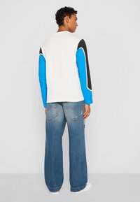 White long-sleeve shirt with blue and black sleeves, paired with wide-leg faded blue jeans. White sneakers complete the outfit.
