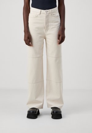 Personne portant un jean large taille haute couleur écrue et des chaussures plateformes noires, debout devant un fond clair uni.