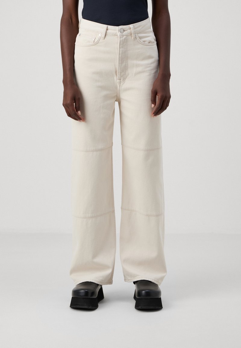 Personne portant un jean large taille haute couleur écrue et des chaussures plateformes noires, debout devant un fond clair uni.