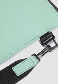 Angolo di una borsa in materiale verde testurizzato con dettaglio della tracolla nera, caratterizzata da una chiusura metallica e cuciture a contrasto lungo i bordi.