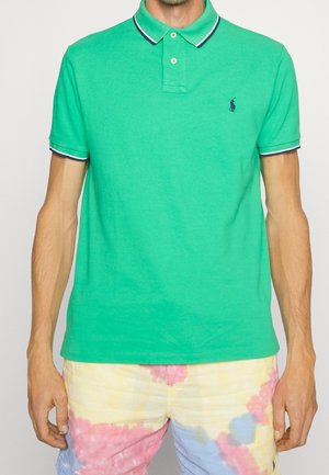 Mężczyzna w zielonej koszulce polo z białym i granatowym wykończeniem oraz wielokolorowych szortach tie-dye na jednolitym tle.