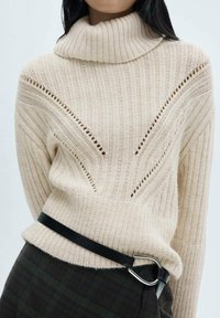 Maglione di colore crema, lavorato a maglia con texture, dotato di un alto colletto a coste e dettagli a taglio, abbinato a una cintura nera in vita.