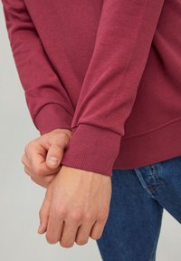 Felpa borgogna con polsini a costine indossata da mani che afferrano le maniche. Il materiale sembra morbido con una texture liscia.