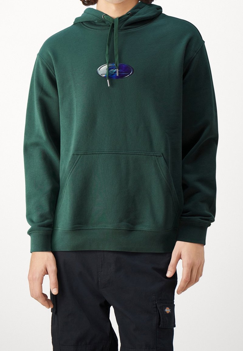 Personne portant un sweat à capuche vert foncé avec poche avant et logo, associé à un pantalon cargo noir sur fond blanc uni.