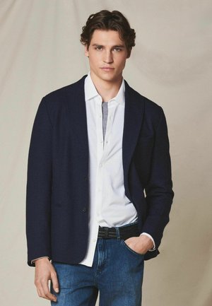 Młody mężczyzna w granatowej marynarce, białej koszuli na guziki i niebieskich dżinsach, stoi z jedną ręką w kieszeni na jednolitym tle.