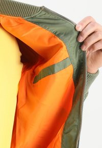 Olivgrön bomberjacka med orange foder, utrustad med ribbat krage och dragkedjeslutning. Synlig ficka på det inre fodret.