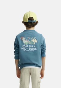 Niño con sudadera azul con ilustraciones de dinosaurios y texto "Roar like a Dino - Silbon Too Cool to Extinct", pantalones beige y gorra amarilla.