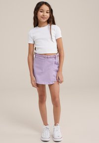 Top corto de manga corta blanco combinado con una falda de mezclilla lila y un cinturón a juego. Zapatillas blancas con acentos rojos completan el atuendo.