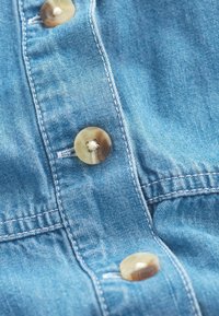 Světle modrá denimbavlna s detaily šití a kulatými, béžovými knoflíky. Detailní textura ukazuje hladký, mírně opotřebovaný vzhled.