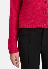 Magenta stickad cardigan med strukturerad yta, med fem rosa knappar framåt, tillsammans med svarta tajta byxor.