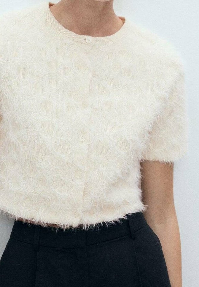 Cardigan court crème et duveteux à manches courtes, avec un col rond et cinq boutons, motif circulaire texturé sur toute la surface.