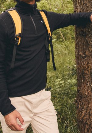 Person trägt schwarzen Fleece mit Reißverschluss und gelbe Rucksackriemen, steht im Freien mit einem Arm, der auf einem Baumstamm ruht.