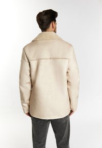 Jachetă bej din blană ecologică cu un guler mare și moale, detalii cusute și croi drept, asortată cu pantaloni gri închis.