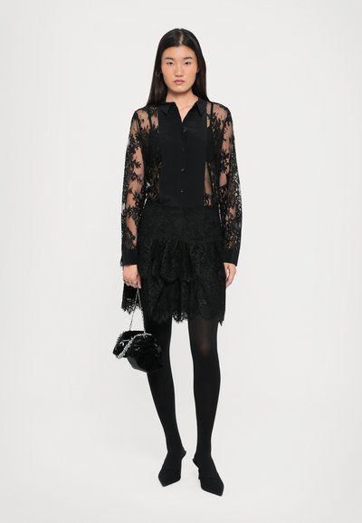 Svart gjennomsiktig blondebluse med lange ermer over en tettsittende svart topp, kombinert med et svart lagdelt blondeskjørt og svarte strømpebukser, holdende en liten svart veske.